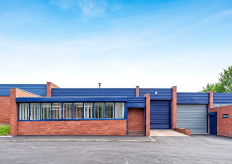 Single-storey red brick industrial building with blue metal roof and cladding, large windows with vertical blinds, a blue roller shutter door, and a paved driveway under a clear sky.