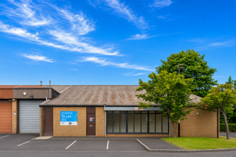 Single-storey commercial building with a To Let sign, large windows, metal roof, empty parking spaces, and trees in front under a blue sky with wispy clouds.