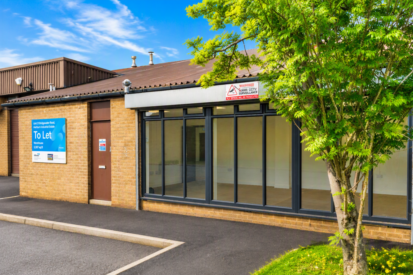 Single-storey commercial building with large front windows, a To Let sign, and a tree in front, situated on a paved car park under a blue sky.