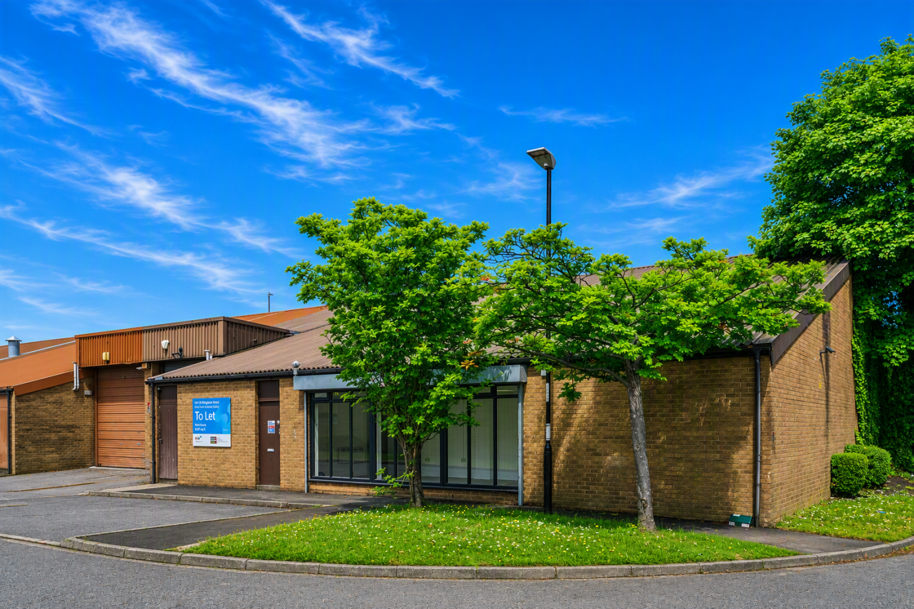 Single-storey brick commercial building with a To Let sign, large windows, garage doors, and two trees in front, under a blue sky with wispy clouds.