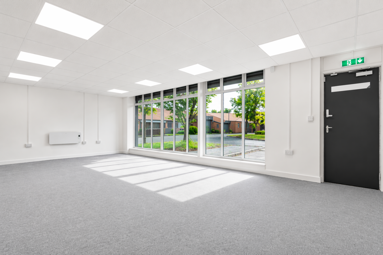 Empty, unfurnished office space with grey carpet, white walls, ceiling lights, large windows letting in natural light, and a black door marked with an emergency exit sign. Greenery can be seen outside.