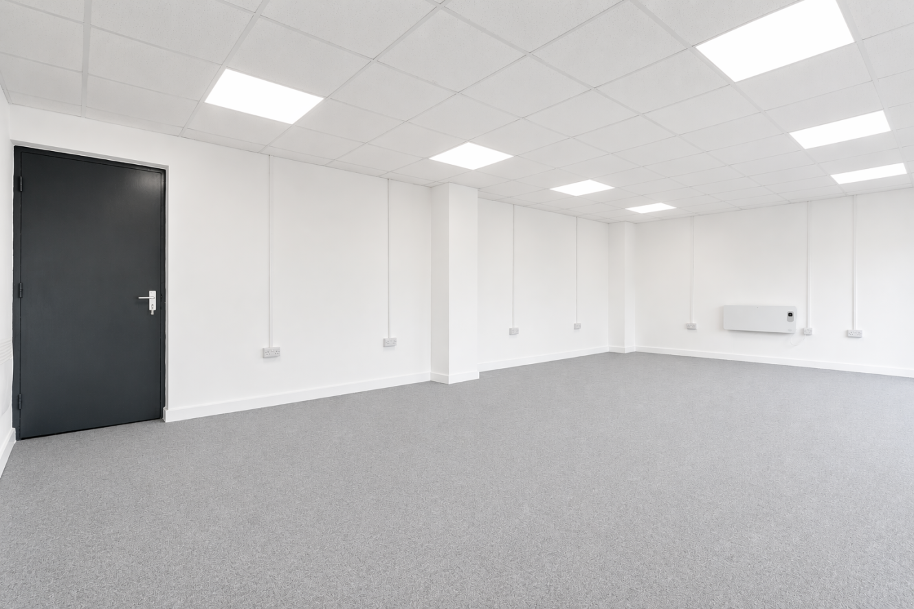 Empty, unfurnished room with white walls, ceiling lights, grey carpet, a closed black door, electrical sockets, and a wall-mounted heater visible.