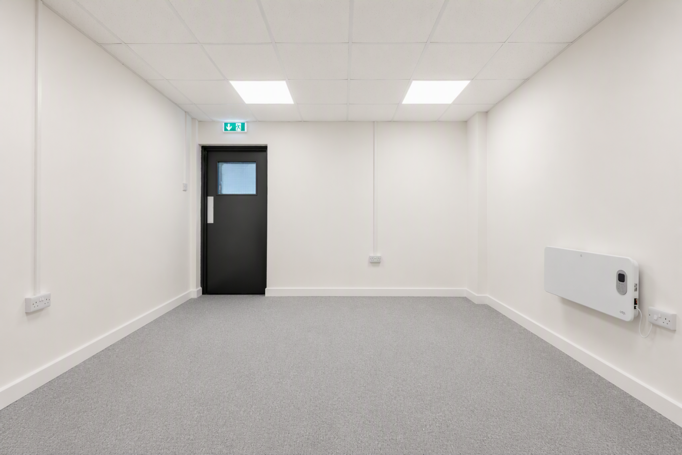 Empty room with white walls, grey carpet, a black door featuring a window, overhead fluorescent lights, and a wall-mounted heater on the right-hand side.