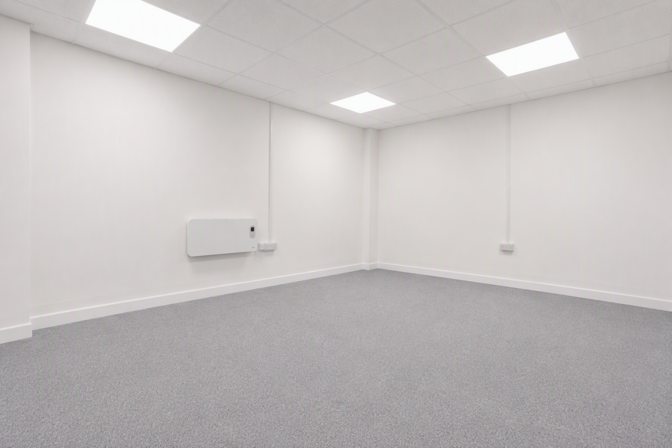 Empty, unfurnished room with white walls, grey carpet, a heater on the wall, electrical sockets, and ceiling lights switched on.