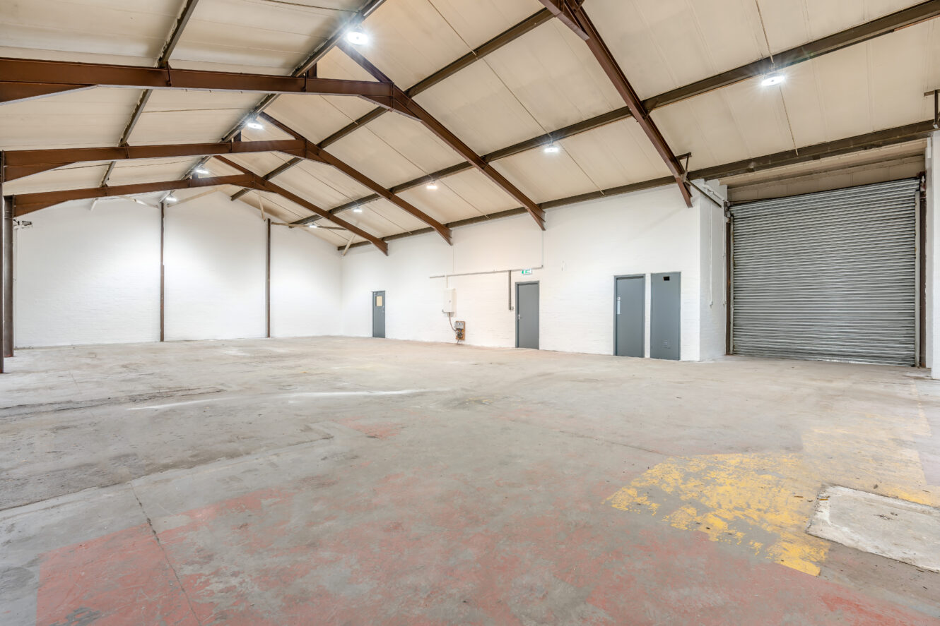 Interior of an empty warehouse with high ceilings, exposed beams, white walls, a concrete floor, multiple doors, and a large metal roller shutter door on the right.