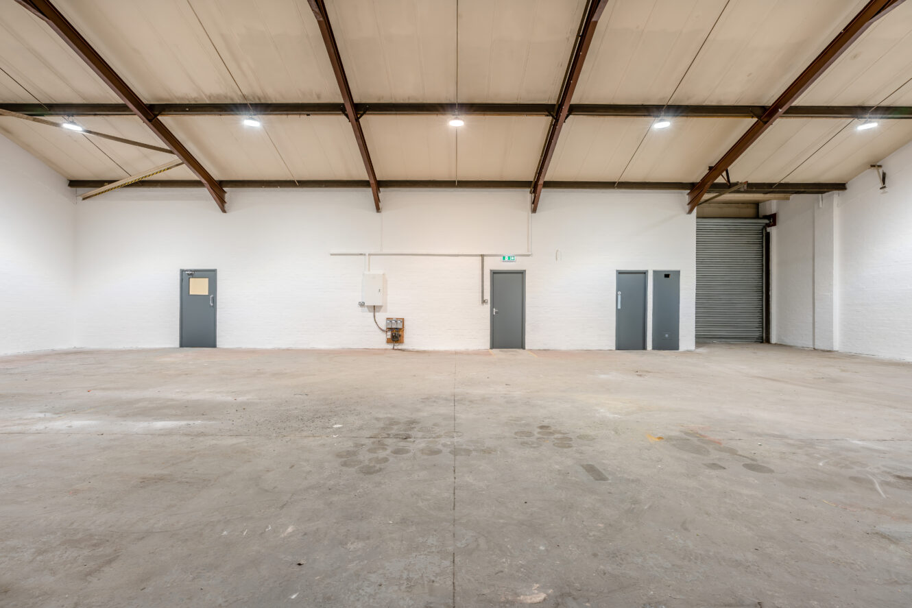 Empty industrial warehouse with a concrete floor, high ceiling, three closed doors, white walls, and a large metal roller shutter door on the right. Fluorescent lights illuminate the space.