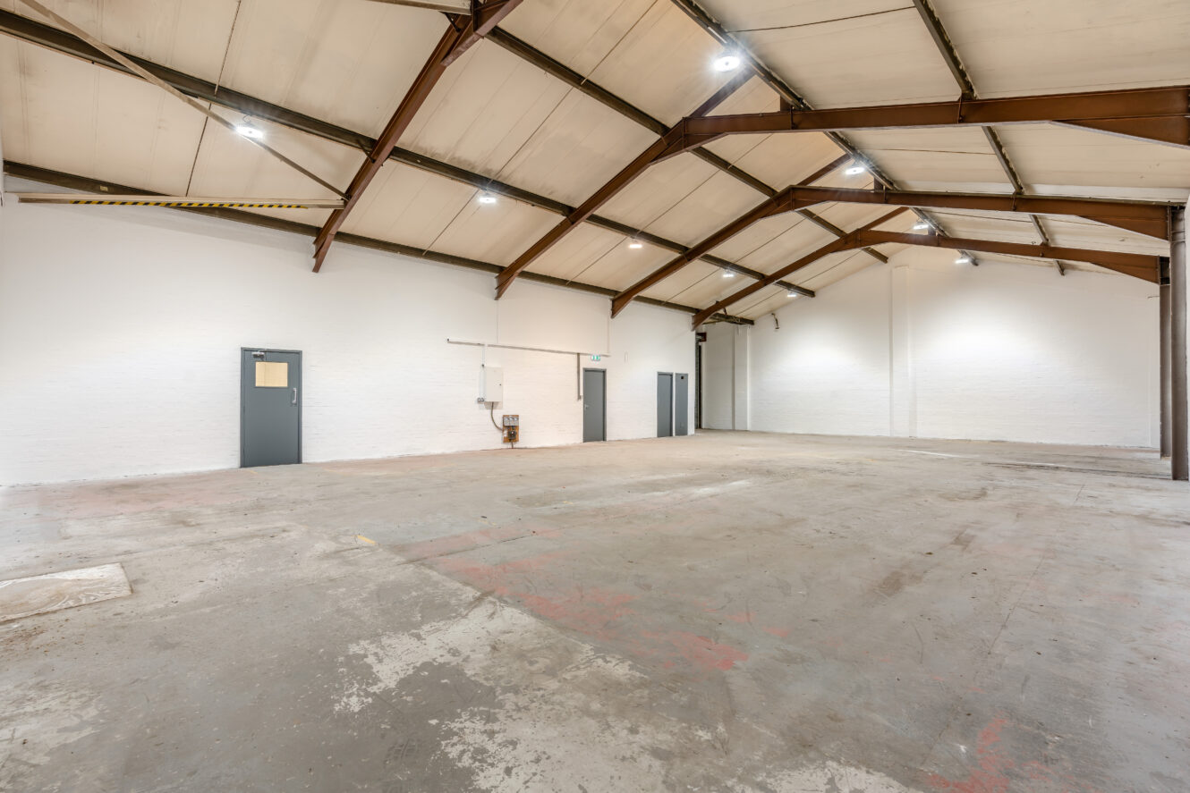 Interior of an empty warehouse featuring a high, beamed ceiling, concrete floor, white walls, and several closed grey doors.