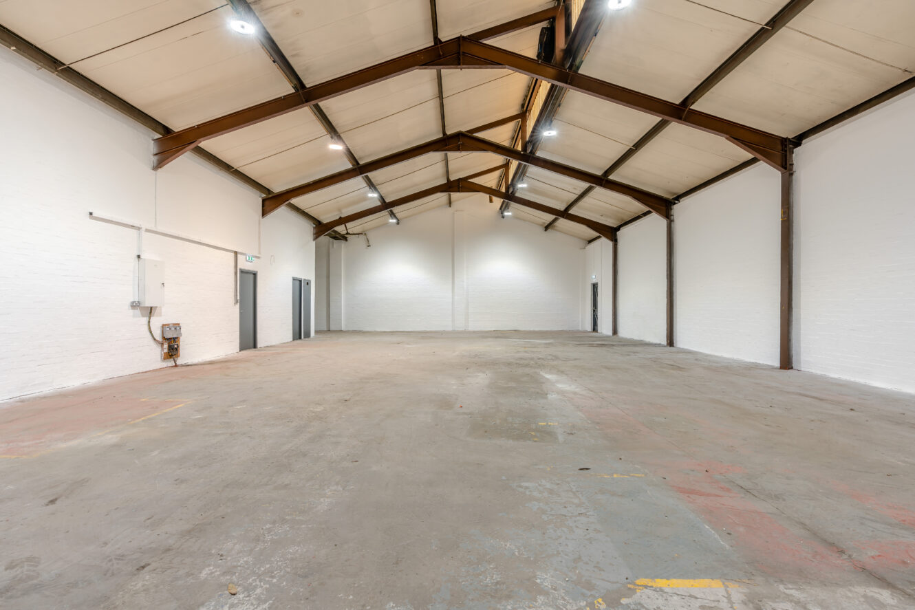 Empty industrial warehouse interior with concrete floor, high ceiling, exposed beams, and white painted brick walls, evenly lit by ceiling lights.