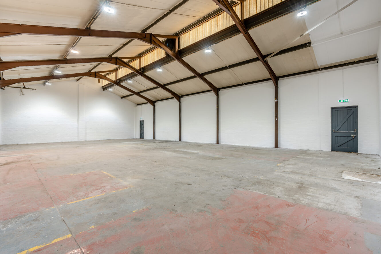 Spacious empty warehouse with a high ceiling, exposed steel beams, concrete floor, white walls, and two grey doors. Overhead lights brightly illuminate the open area.