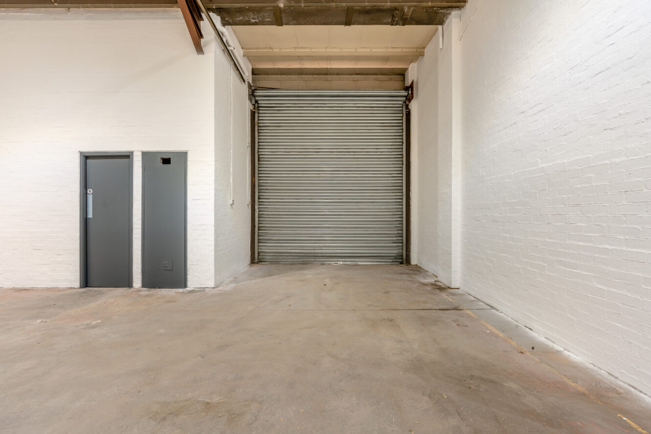 Empty industrial space with a large metal roller shutter, white brick walls, and a grey door to the left.