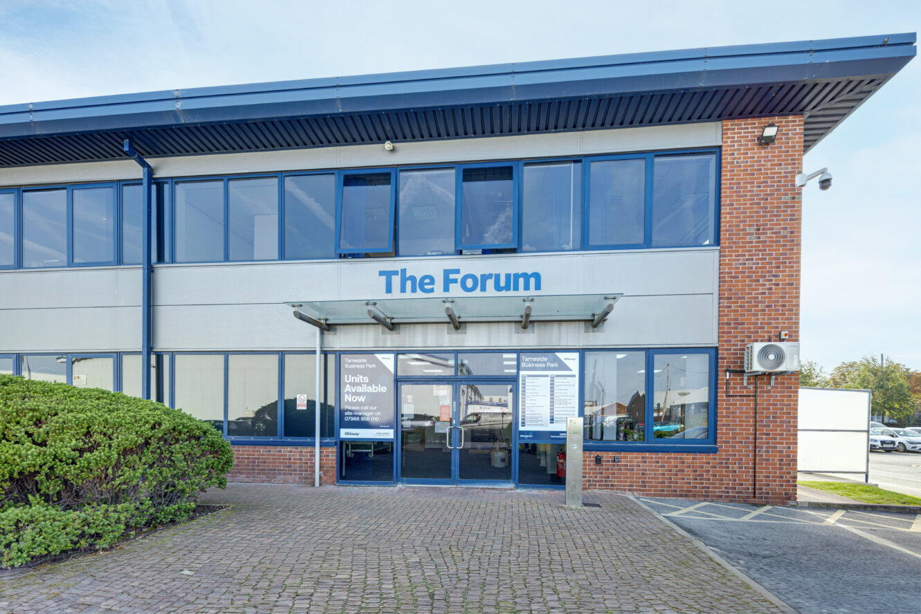 Two-storey commercial building with large windows and a sign reading The Forum above glass entrance doors. Shrubs and paved walkway are in the foreground.