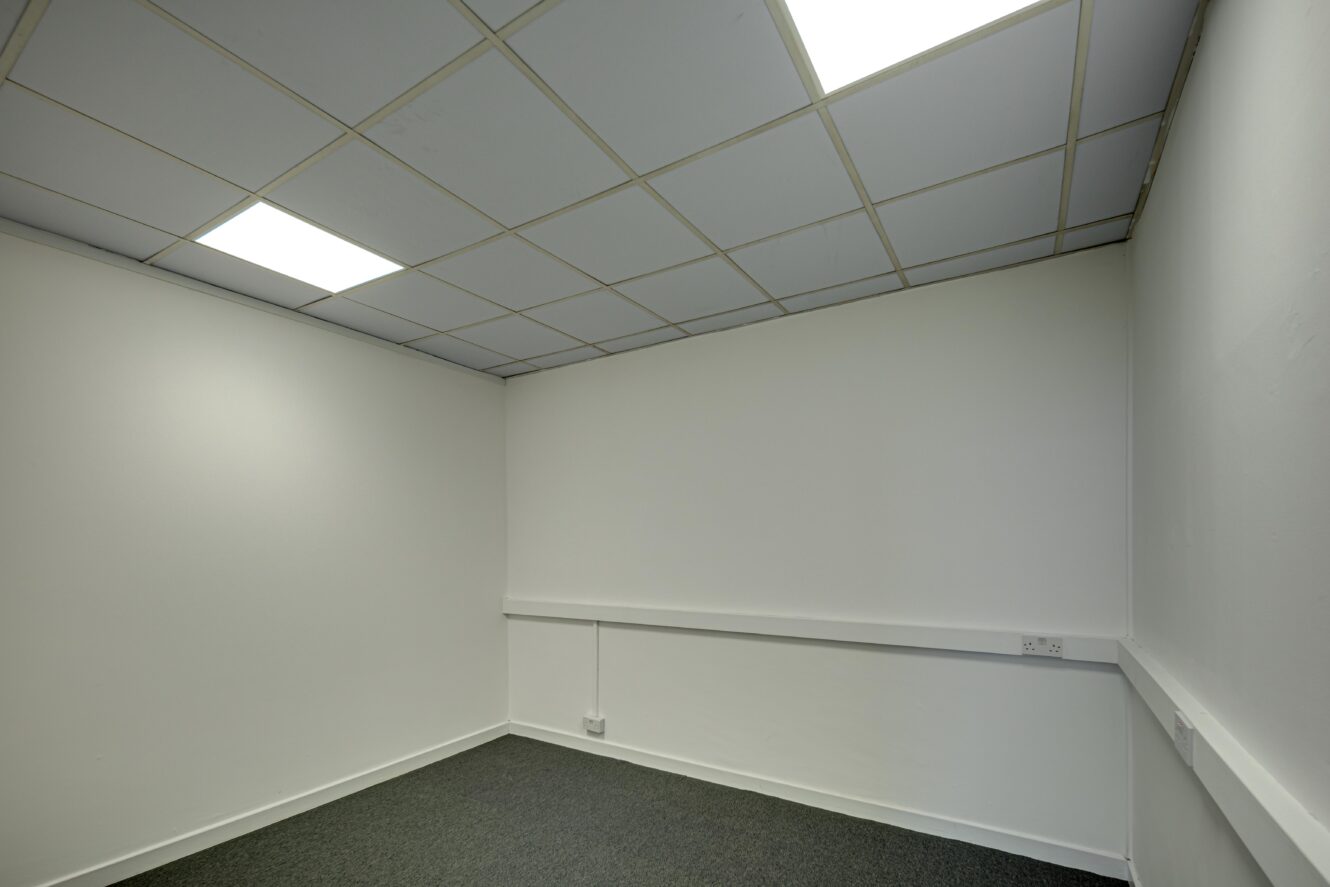 Empty office room with white walls, grey carpet, suspended ceiling featuring fluorescent lights, and electrical sockets positioned along one wall.