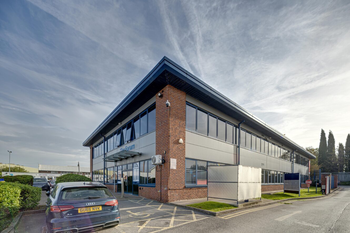 Modern two-storey office building with brick and metal exterior, large windows, and parked cars in front, photographed on a partly cloudy day.