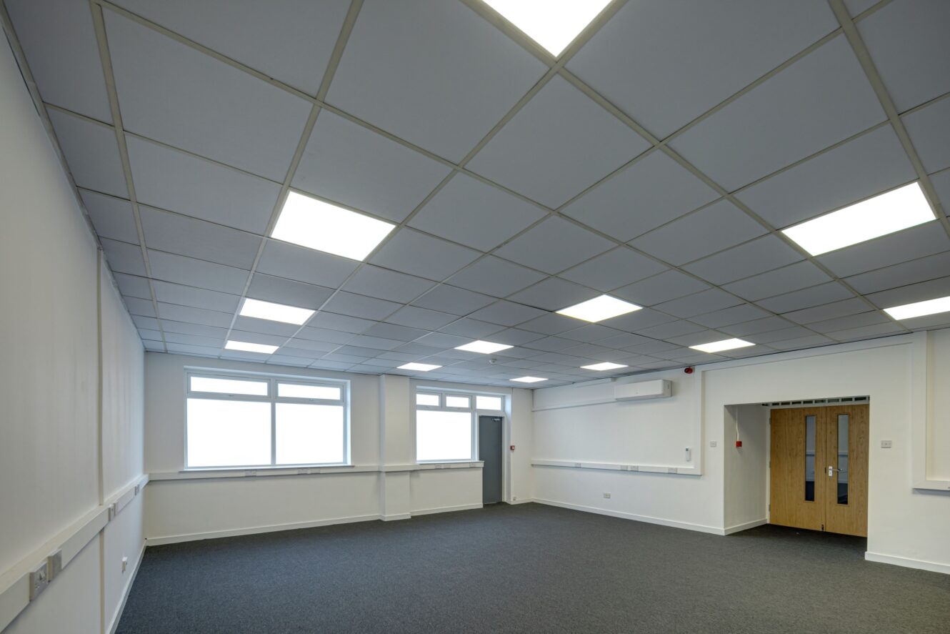 Empty office room with grey carpet, white walls, ceiling grid featuring fluorescent lights, two windows, and wooden double doors.