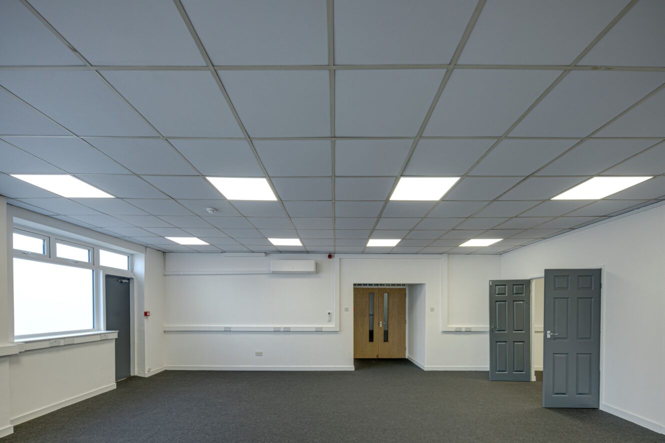 Empty office room with white walls, grey carpet, ceiling lights, closed grey doors on the right, and double wooden doors at the back. A large window on the left side allows natural light into the space.