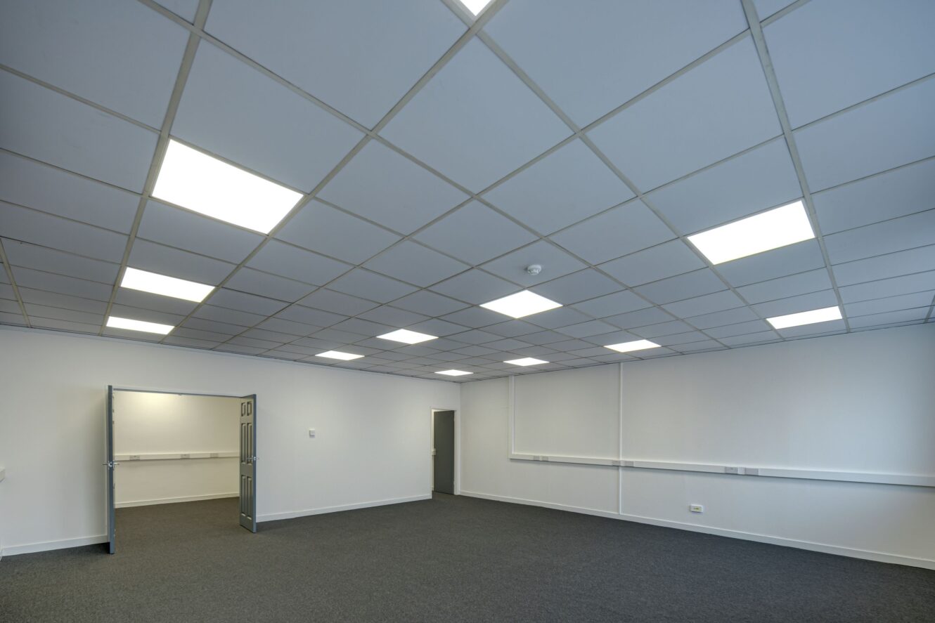 Empty office room with grey carpet, white walls, suspended ceiling tiles, and fluorescent ceiling lights. An open door leads to a similar adjoining room.