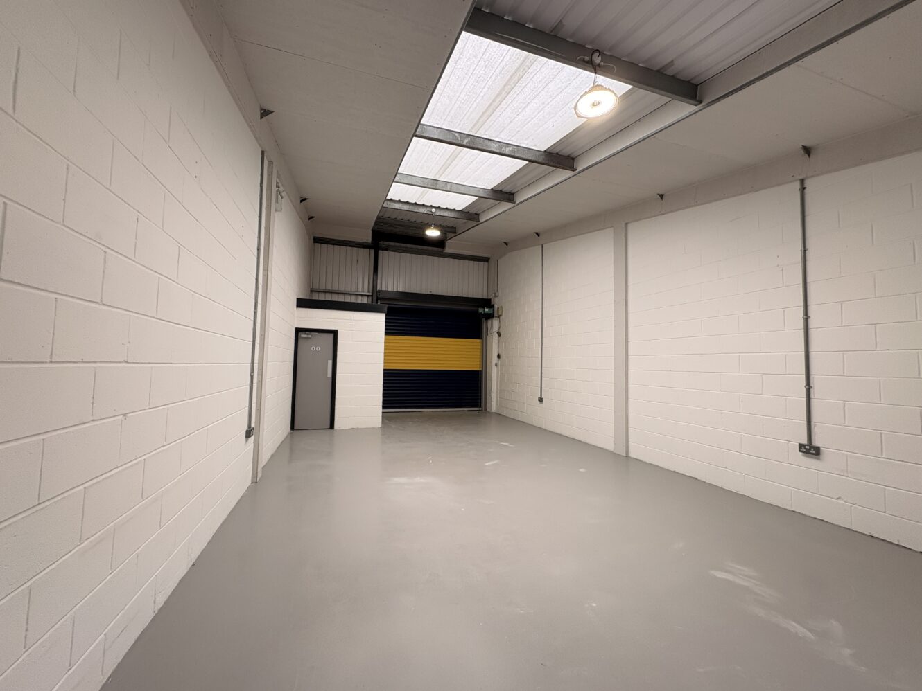 Empty industrial unit with white brick walls, a grey floor, skylights, fluorescent lights, a metal door on the left, and a large blue and yellow roller shutter at the far end.
