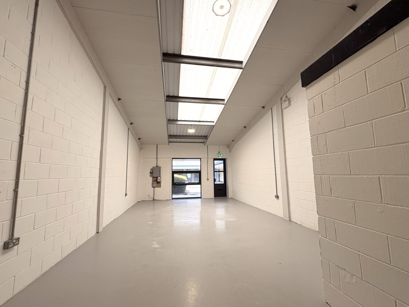 Empty industrial unit with white brick walls, grey floor, exposed wiring, and skylights. A door and window are visible at the far end, allowing natural light into the space.