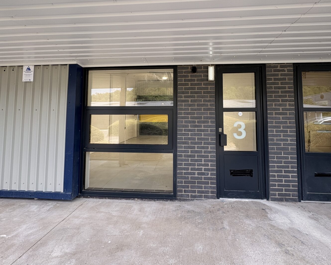 Exterior view of a building with a glass window, a door labelled 3, and brick walls under a white overhang.