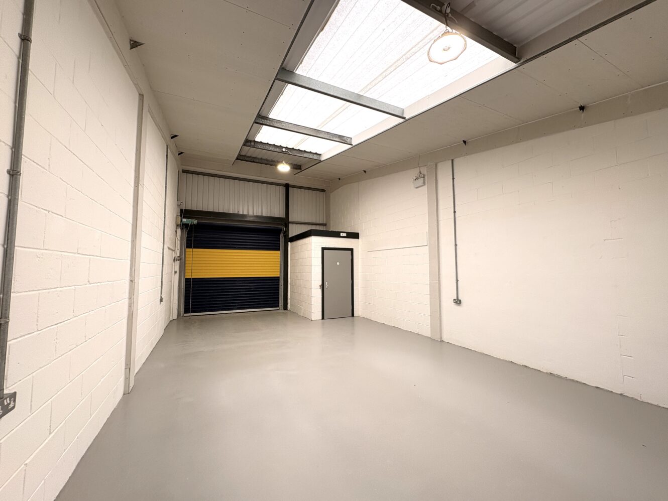 Empty industrial unit with white brick walls, grey concrete floor, skylights, exposed pipes, a grey door, and a blue-and-yellow roller shutter at the far end.