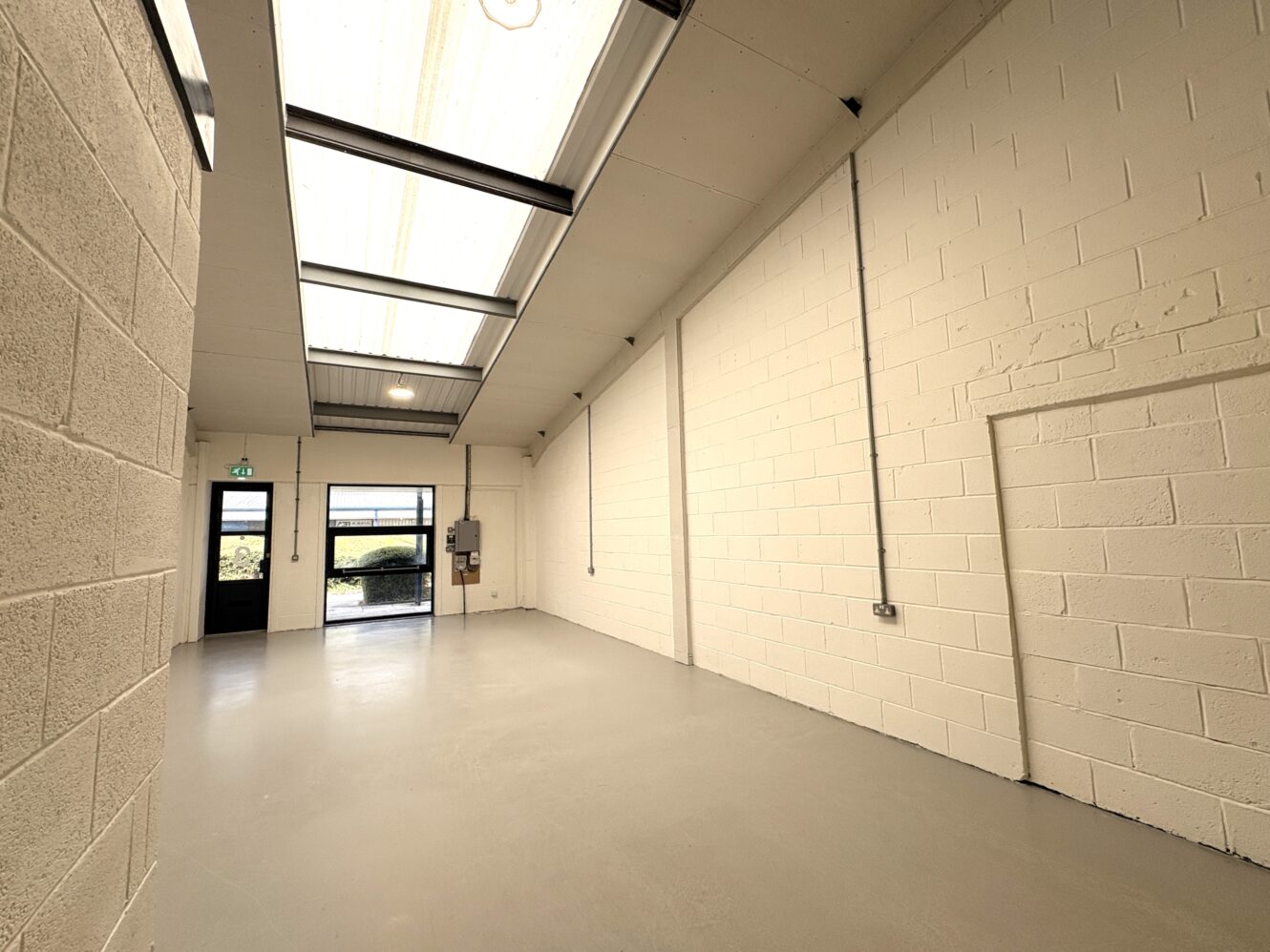 Empty industrial unit with cream brick walls, grey concrete floor, high ceiling, skylights, a wall-mounted electrical box, and a black-framed door leading outside.
