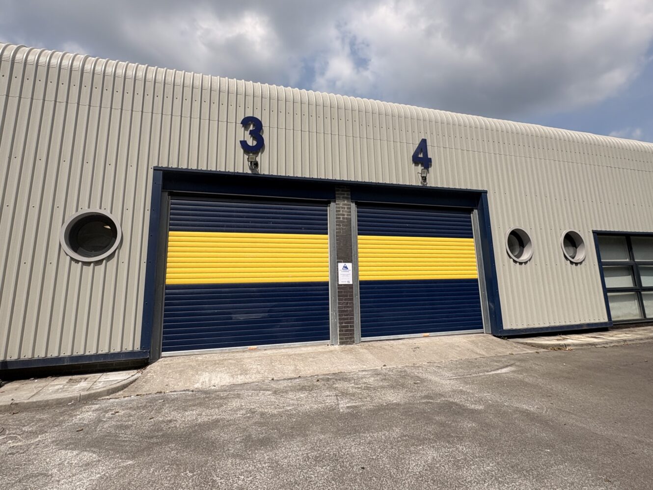 Two large garage doors numbered 3 and 4, marked with blue and yellow stripes on a grey industrial building beneath a cloudy sky.
