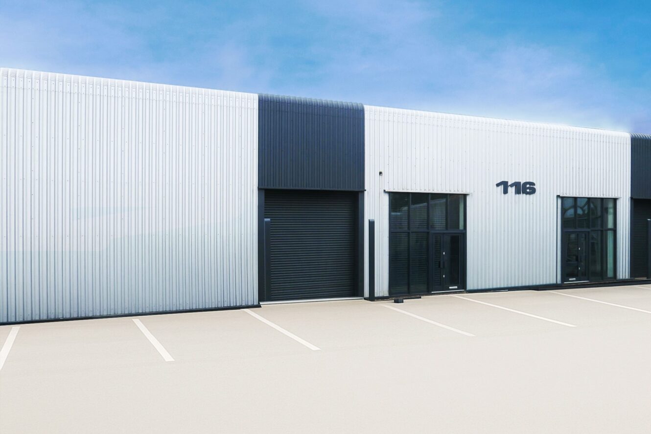 A modern industrial building with metal cladding, tinted windows, a black garage door, and the number 116 above the entrance; the empty car park has white lines.
