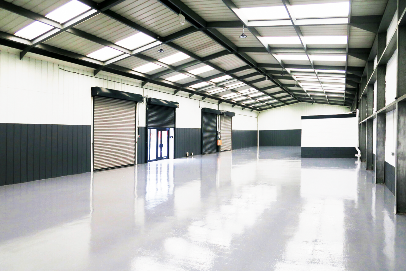 Large empty warehouse with shiny grey floors, high ceiling, and several roller shutters along one side. Skylights and lights are visible on the roof.