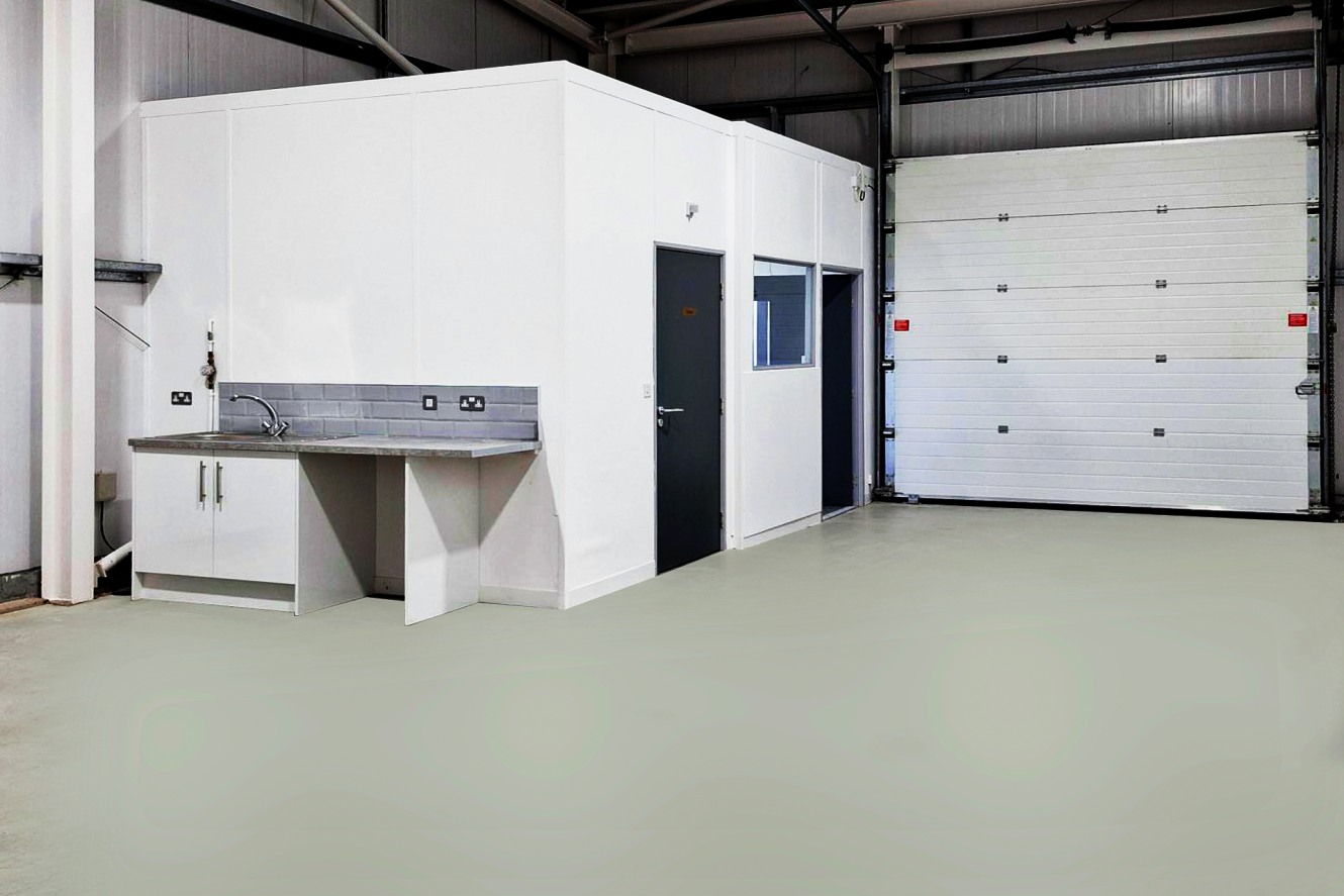 A clean, empty warehouse with a small white office unit, a sink with cupboards, and a large closed roller shutter door.