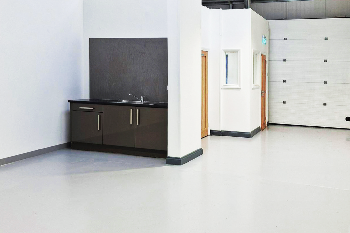 A small kitchenette with a sink and cupboards is set against a partial wall in a large, empty, industrial-style room with white walls and grey flooring.
