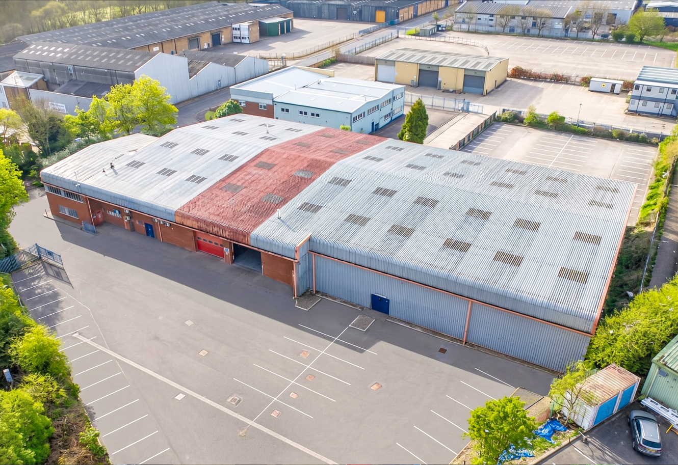 Aerial view of a large industrial warehouse with a worn and rusted roof, surrounded by empty car parks and nearby industrial buildings.