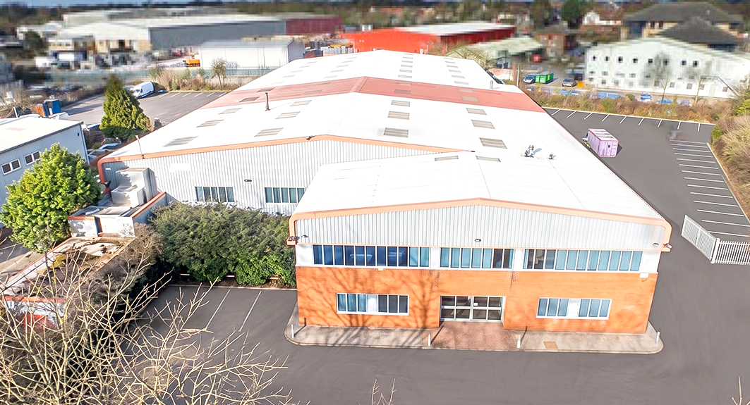 A large industrial warehouse with a brick and metal exterior, surrounded by a tarmac car park, seen from an elevated angle.