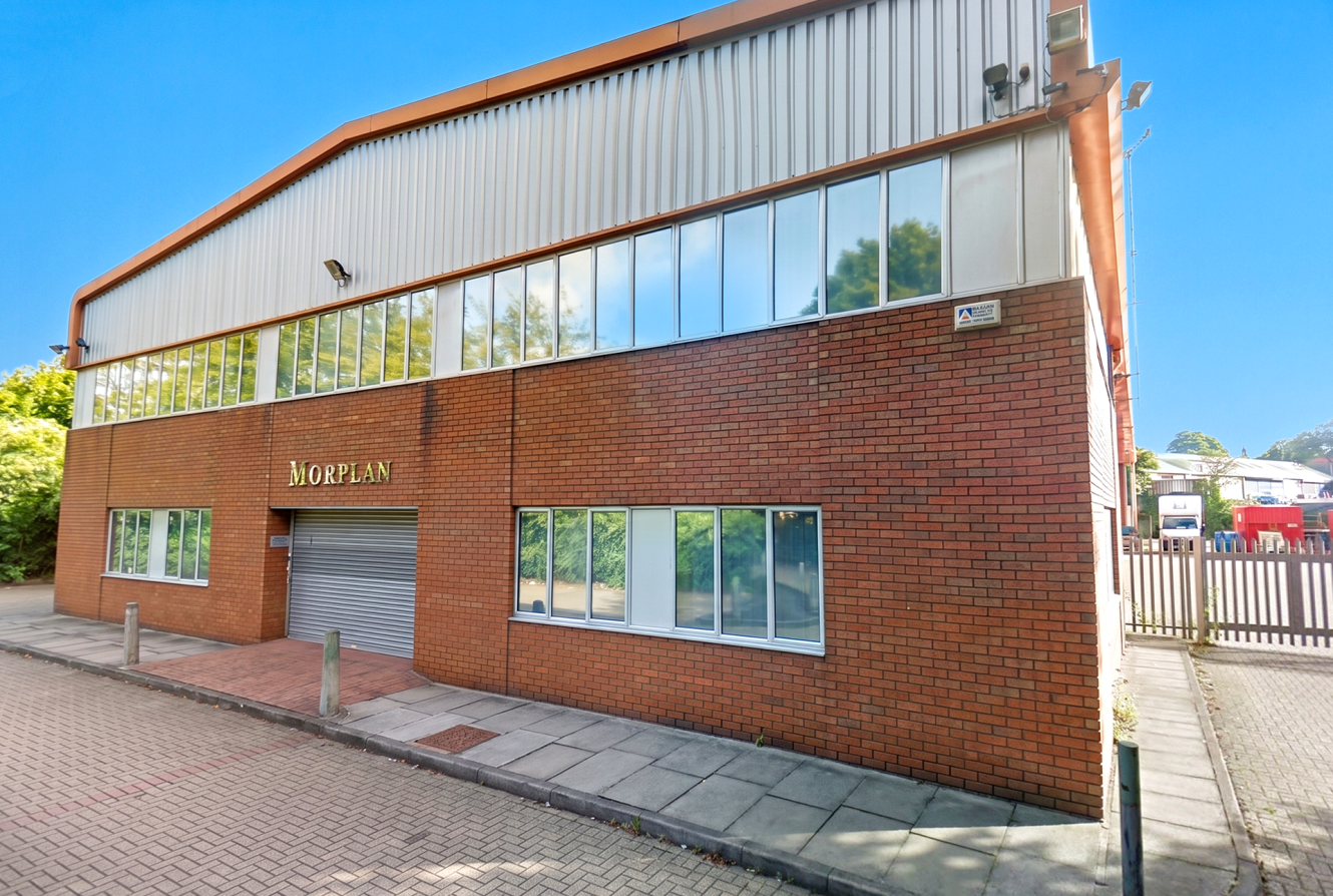 A two-storey brick and metal commercial building labelled Moreplan with large windows and a closed rolling door on a sunny day.
