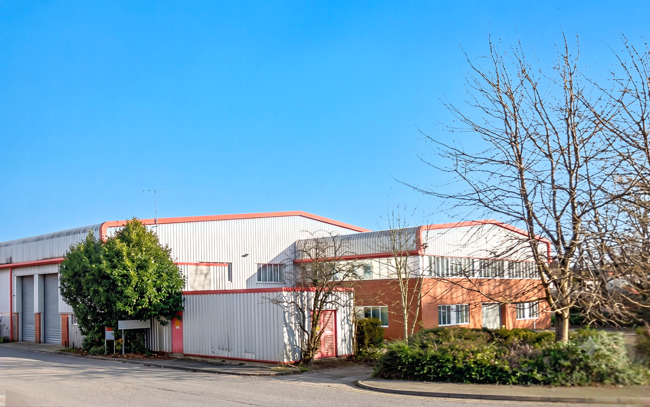 A modern industrial warehouse with red trim, brick and metal walls, large windows, and trees in front, under a clear blue sky.