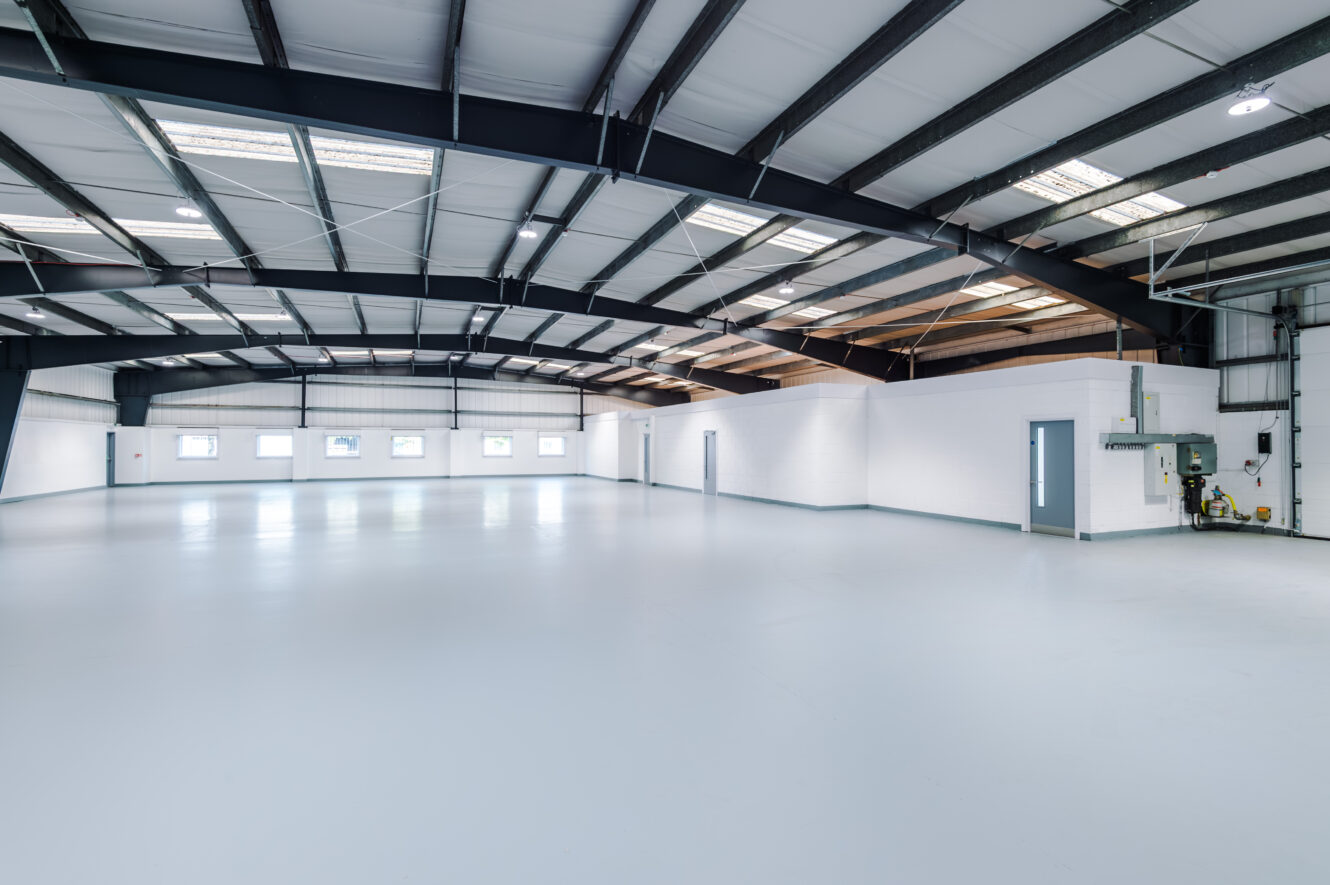 Spacious empty warehouse with white floors and walls, grey doors, exposed beams, and windows along one side.