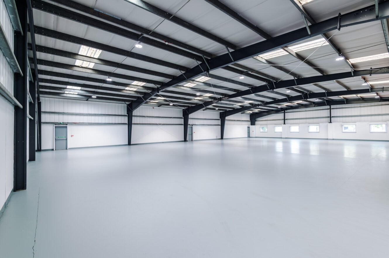 Spacious empty warehouse with white walls, high ceiling, exposed steel beams, and several windows along the side.