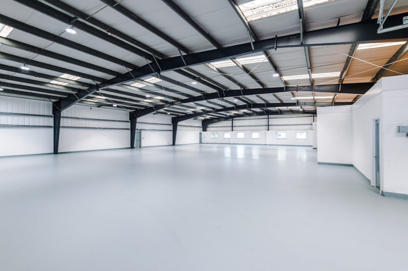 Large empty industrial warehouse featuring white floors, metal beams, windows along one wall, and skylights in the ceiling.