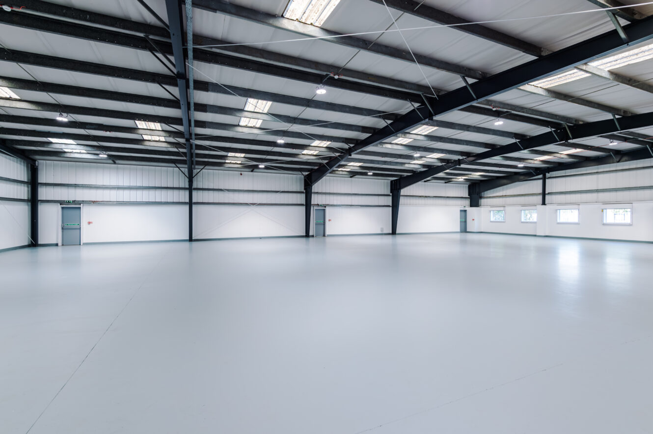 Spacious, empty industrial warehouse with white walls, grey floor, high ceiling with metal beams, and several small windows and doors along the sides.