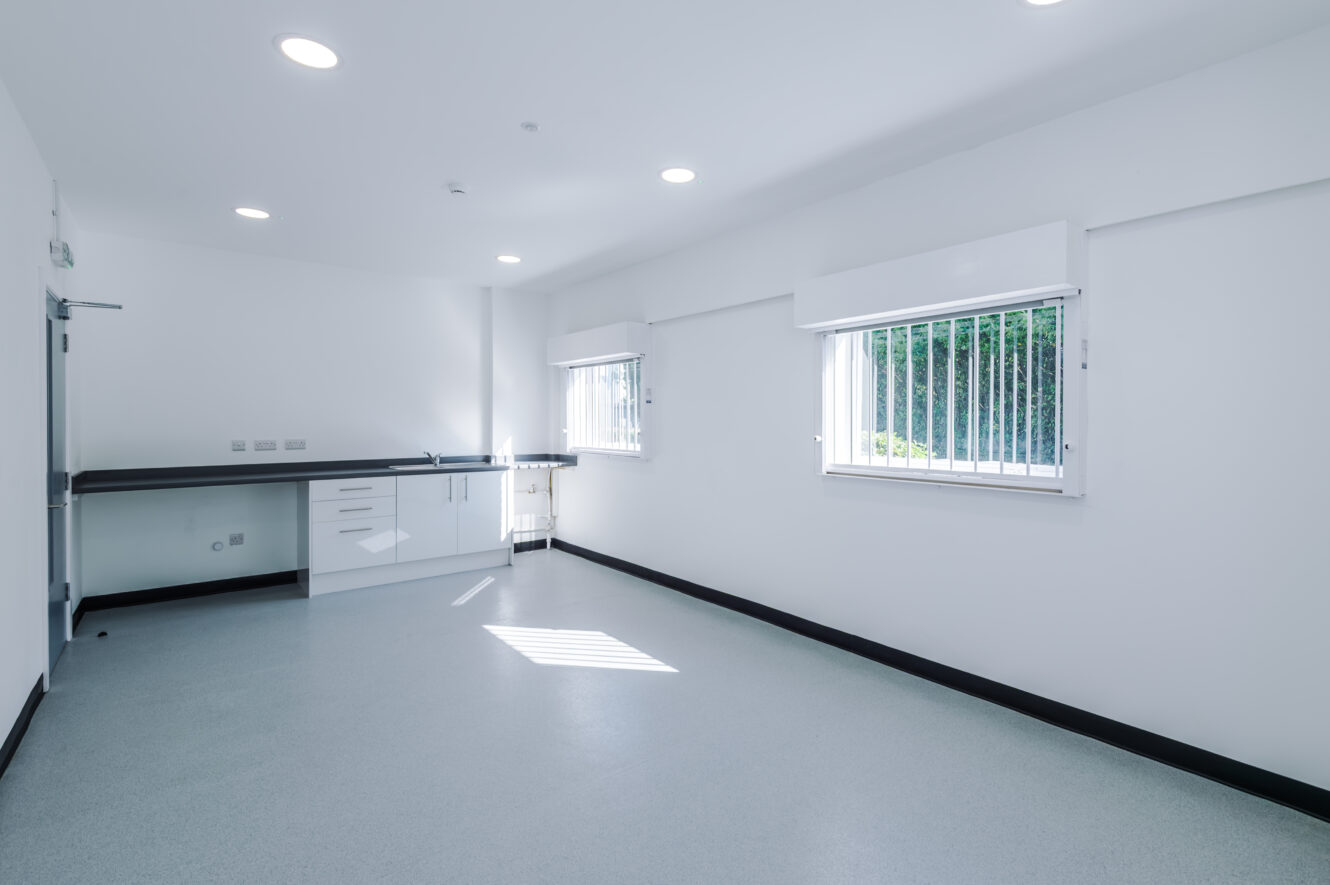 Empty, bright clinical room with white walls, two barred windows, overhead lights, and a worktop with drawers and cupboards along one side.