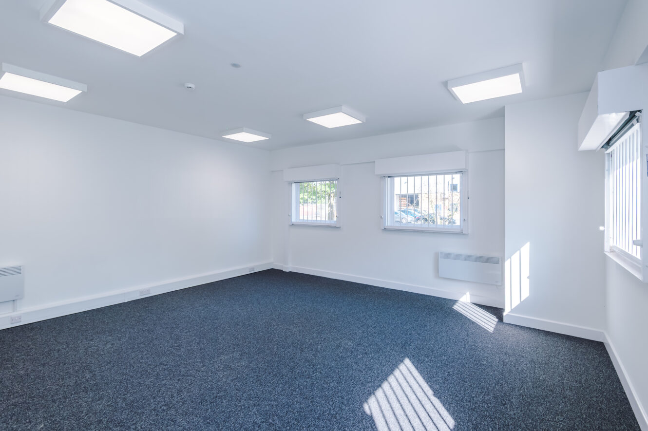 Empty, unfurnished room with white walls, three barred windows, blue carpet, and ceiling lights. Sunlight streams in through one of the windows.