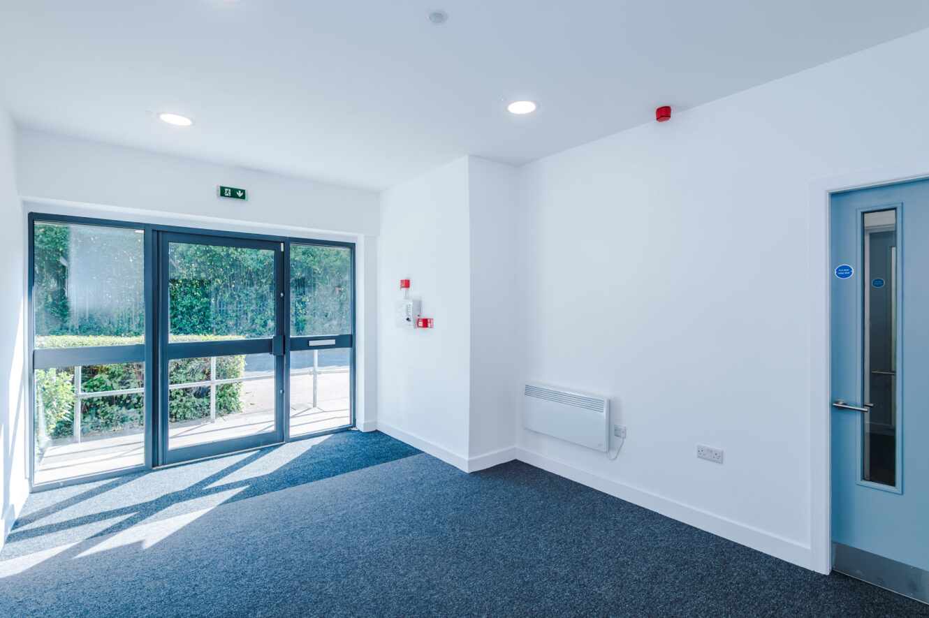 Bright, empty room with blue carpet, white walls, glass double doors leading outside, a blue internal door, and a small wall heater. Emergency exit signs and fire safety equipment are visible.
