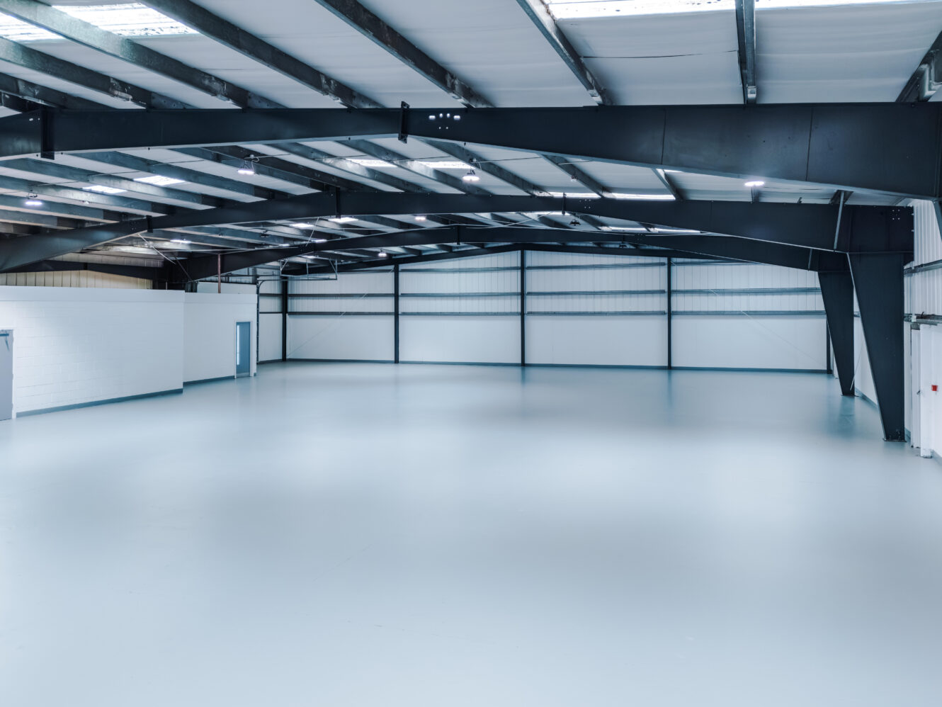 Large empty warehouse interior with white floors, metal beams, high ceiling, and white walls; several doors are visible along the side and back.