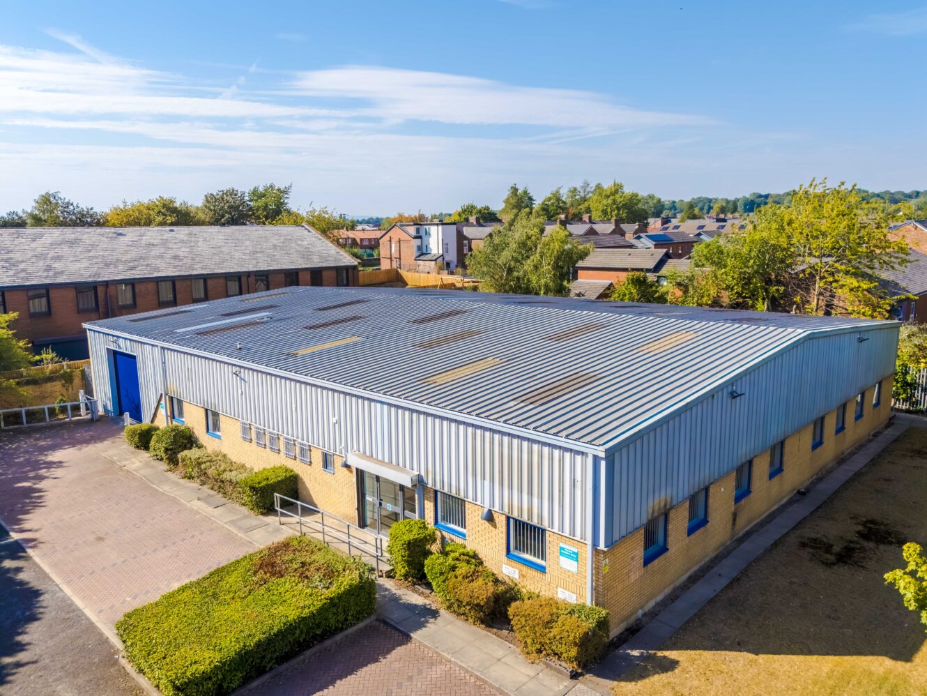 Single-storey industrial warehouse with a metal roof, brick walls, small windows, and surrounding bushes, set in a business park area with neighbouring buildings and a clear sky.