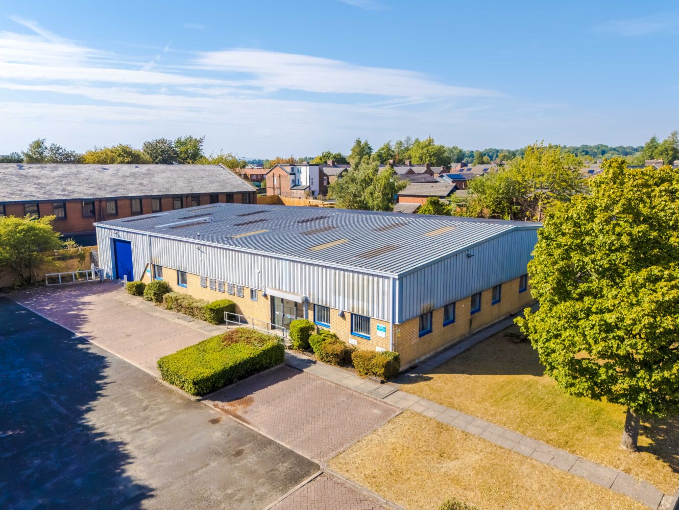 Single-storey industrial warehouse building with a metal roof, brick walls, blue trim, and a car park, surrounded by trees and other buildings under a clear sky.