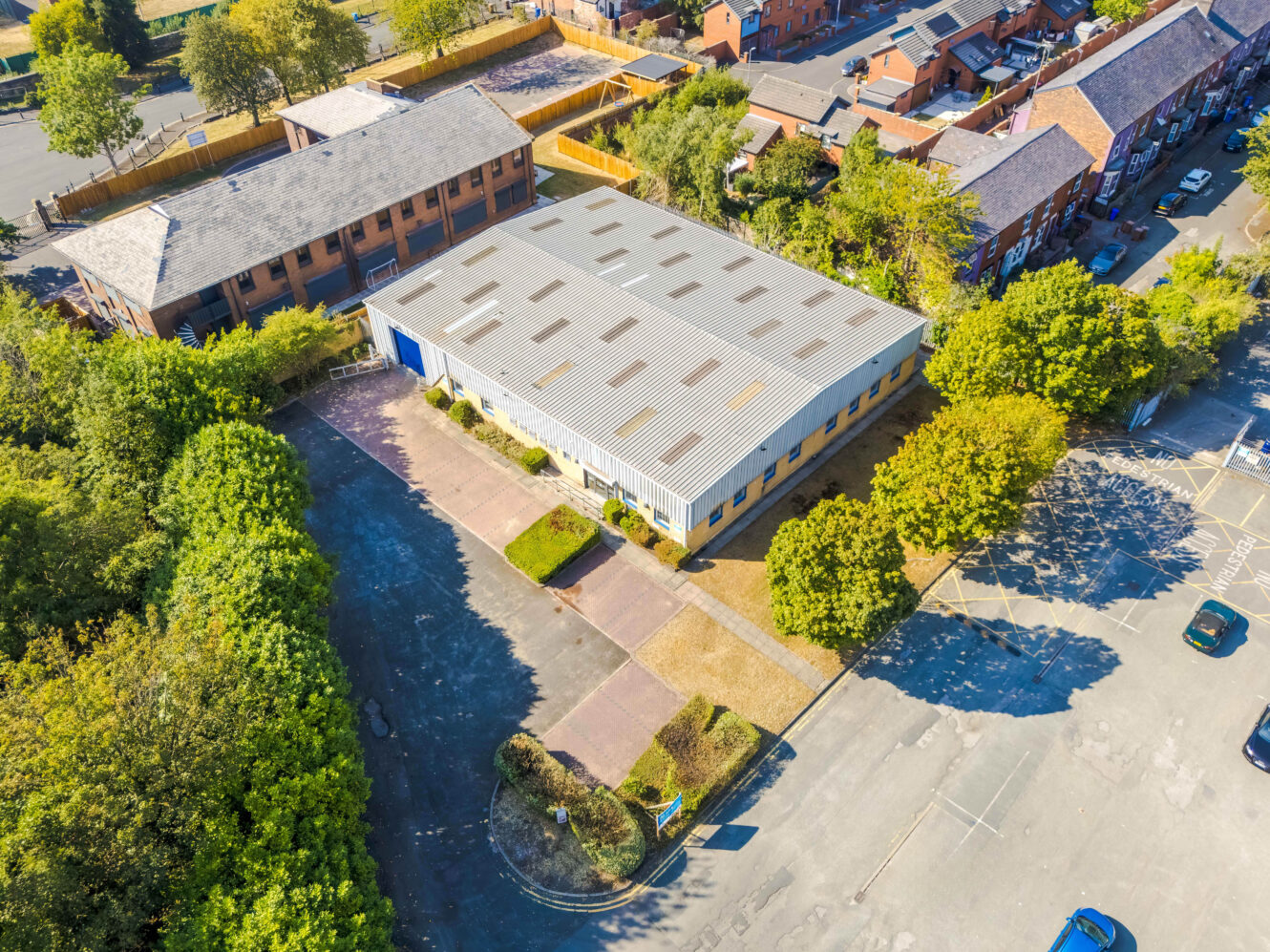 Aerial view of a single-storey industrial building with a metal roof, surrounded by trees, parking spaces, and nearby residential and commercial buildings.