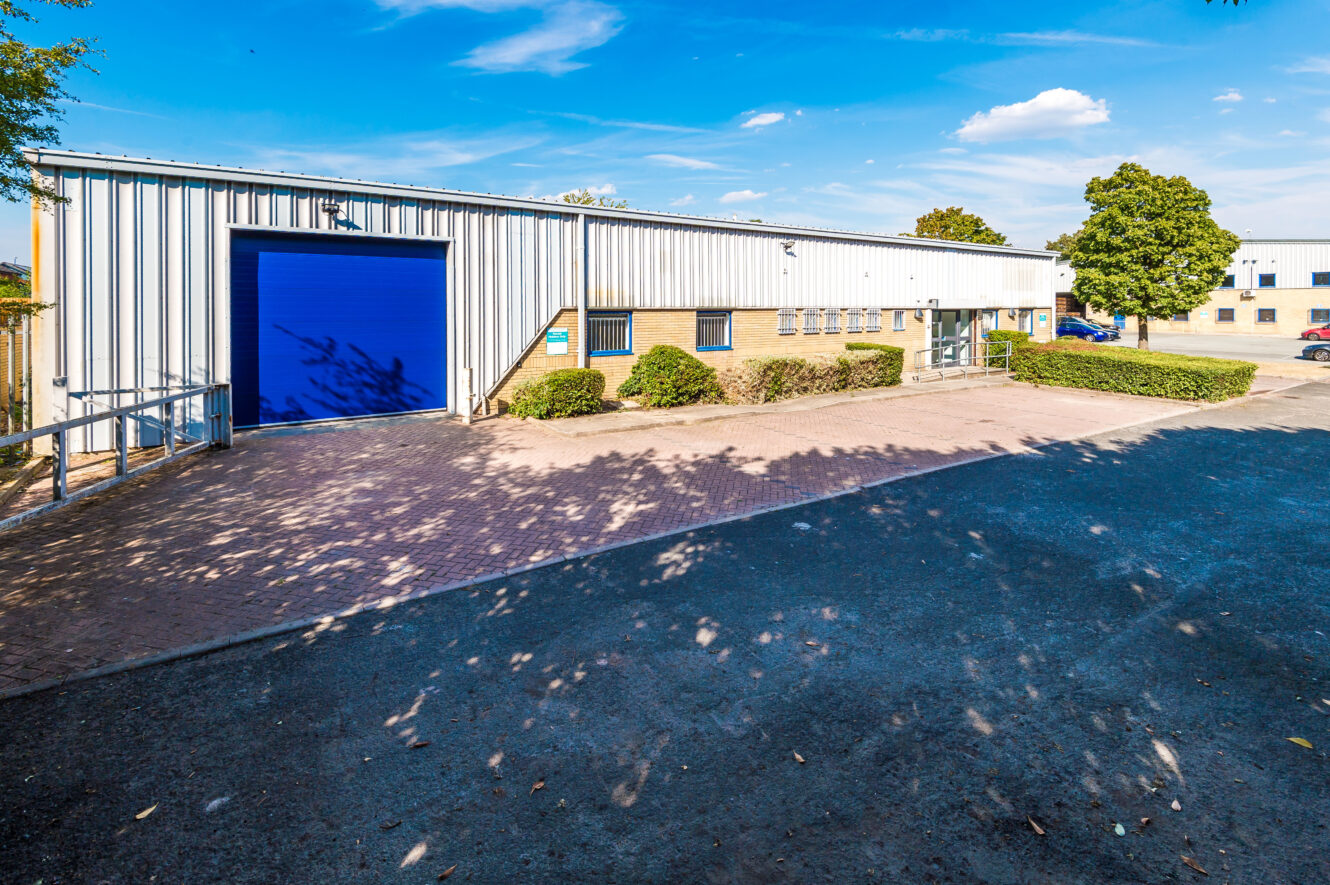 A single-storey industrial building with a blue loading bay door, brick paving, small bushes, and a car park under a clear blue sky.