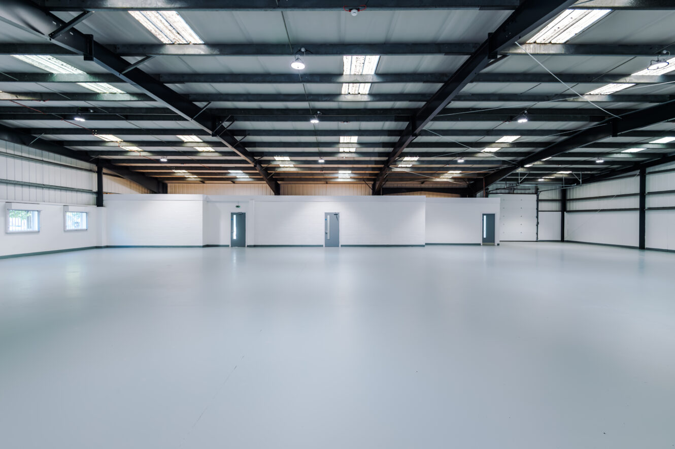 Large, empty warehouse with white walls, grey doors, high ceilings, exposed beams, and overhead fluorescent lighting.