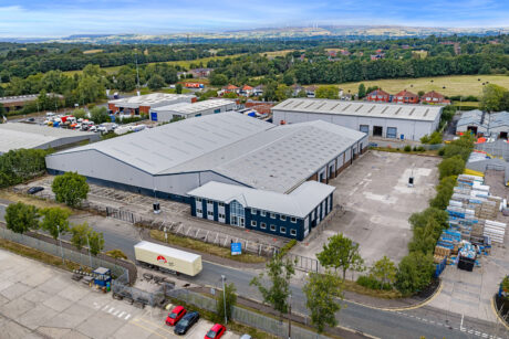 Aerial view of a large industrial warehouse complex with parking areas, shipping containers, and nearby greenery under a partly cloudy sky.