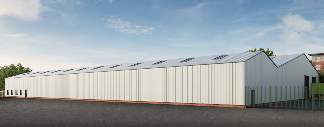 Large white industrial warehouse with a zigzag roof, rooflights, and a gated entrance, situated on a tarmac lot under a clear sky.