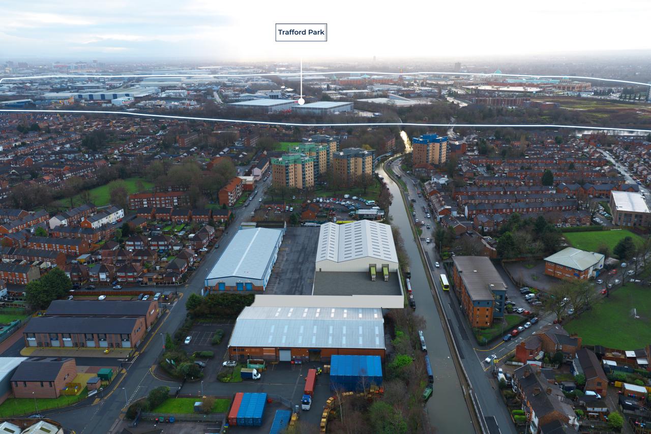 Aerial view of an urban area featuring industrial buildings in the foreground, residential houses, a canal, and a label indicating Trafford Park in the distance.
